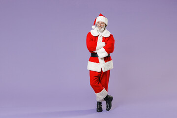 Full length portrait smiling Santa Claus man in Christmas hat red suit coat glasses holding hands crossed looking aside isolated on violet background. Happy New Year celebration merry holiday concept.