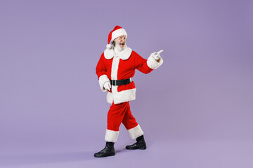 Full length side view portrait of excited Santa Claus man in Christmas hat red coat suit pointing index finger aside isolated on violet background. Happy New Year celebration merry holiday concept.
