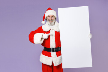 Smiling Santa Claus man in Christmas hat red coat suit glasses pointing index finger on big white empty blank billboard isolated on violet background. Happy New Year celebration merry holiday concept.