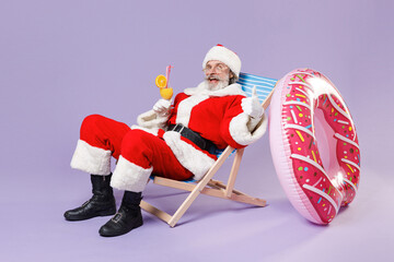 Funny Santa Claus man in Christmas hat suit coat glasses sit on deck chair hold glass with cocktail showing thumb up isolated on violet background. Happy New Year celebration merry holiday concept. - Powered by Adobe