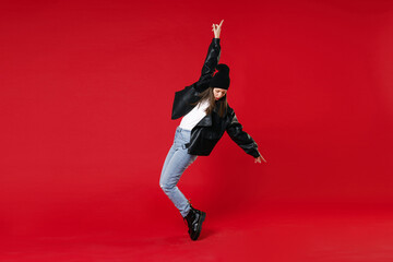 Fototapeta na wymiar Full length of funny young brunette woman 20s in black leather jacket white t-shirt hat pointing index finger up dancing standing on toes isolated on bright red colour background studio portrait.