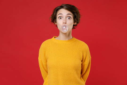 Amazed Funny Pretty Cute Young Brunette Woman 20s Wearing Basic Yellow Sweater Standing Chewing, Blowing Bubble Gum Balloon Looking Camera Isolated On Bright Red Colour Background, Studio Portrait.