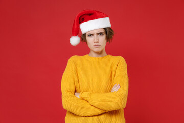 Displeased young brunette Santa woman wearing casual yellow sweater Christmas hat holding hands crossed isolated on red background studio portrait. Happy New Year celebration merry holiday concept.