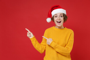 Excited young brunette Santa woman in casual yellow sweater Christmas hat pointing index fingers...