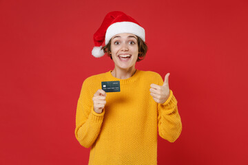 Excited young Santa woman in casual yellow sweater Christmas hat hold credit bank card showing thumb up isolated on red background studio portrait. Happy New Year celebration merry holiday concept.
