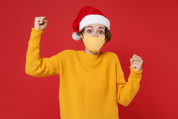 Excited Santa woman in Christmas hat face mask safe from coronavirus virus covid-19 clenching fists doing winner gesture isolated on red background. Happy New Year celebration merry holiday concept.