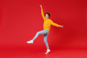Fototapeta na wymiar Full length side view of cheerful excited beautiful young brunette woman 20s wearing basic yellow sweater dancing rising spreading hands legs isolated on bright red colour background studio portrait.