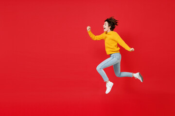 Full length side view of excited surprised cheerful funny young brunette woman 20s wearing basic casual yellow sweater jumping like running isolated on bright red colour background studio portrait.