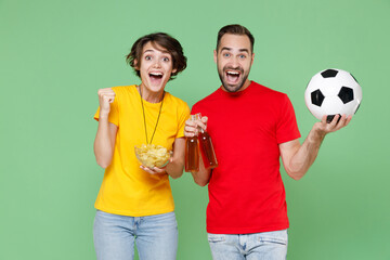 Excited happy young couple friends sport family woman man football fans in t-shirts cheer up support favorite team with soccer ball hold beer bottles chips clenching fist isolated on green background.