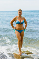 Obraz premium A young blonde woman in a blue swimsuit stands in the water on the stones, on the seashore.