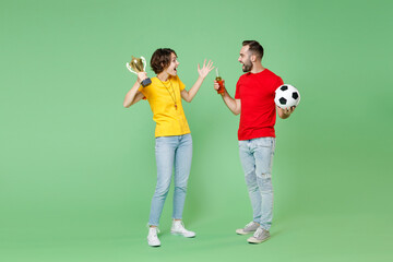 Fototapeta na wymiar Full length of shocked couple friends sport family woman man football fans in t-shirts cheer up support favorite team with soccer ball hold beer bottle winner cup isolated on green background studio.
