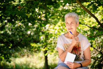 Naklejka na ściany i meble Smiling senior hiker lady thinks and takes notes in the forest. nature concept.