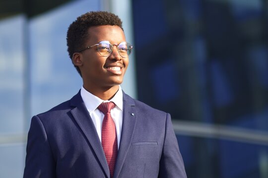 Portrait of handsome happy cheerful positive businessman in formal suit, tie and glasses. Black African Afro American successful man standing outdoors business building, smiling, looking into distance