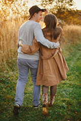 Happy stylish couple hugging and walking in autumn meadow in warm sunset light
