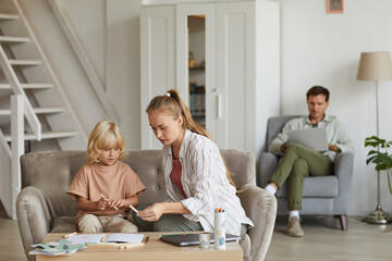 Obraz premium Mother teaching her son at the table in the room with father working on laptop in the background