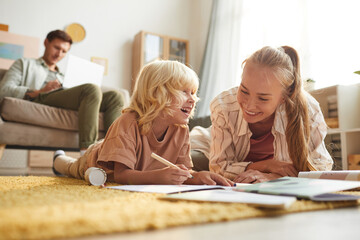 Fototapeta premium Happy mother lying on the floor together with her son and they drawing with father sitting on sofa in the background