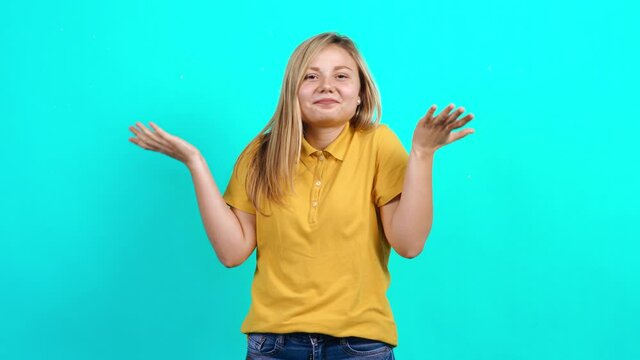 The confused young woman points with her right finger. Beautiful Caucasian woman in yellow T-shirt and long blond hair standing on blue background. A little fat teenager. Advertising concept.