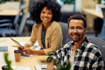 Two young cheerful male and female office workers sitting at desk , smiling at camera while working...