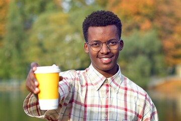 Portrait of young happy positive man in shirt and glasses holding out a plastic cup of hot drink tea or coffee, smiling, looking at camera. Black African Afro American guy in golden autumn park. 