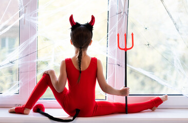 Little girl gymnast in costume of little devil doing stretching exercise on window sill