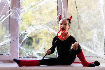 Little girl gymnast in costume of little devil playing about and stretching on window sill
