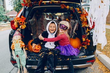 Trick or trunk. Children celebrating Halloween in trunk of car. Boy and girl with red pumpkins celebrating traditional October holiday outdoors. Social distance and safe alternative celebration. - 385367268