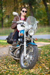 Female motorcycle rider sitting on a classic motorbike, woman with helmet in hands. Portrait in autumn park