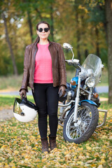 Female motorcycle rider standing next to a motorbike, woman holding helmet in hand. Full-length portrait in autumn park