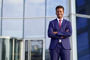 Fototapeta premium Portrait of serious confident successful black African man, Afro American young businessman in formal suit with tie standing outdoors business office with his hands crossed, looking at camera