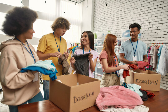 Happy young volunteers group sorting, packing clothes in cardboard boxes, Diverse team working together on donation project in charitable organization office