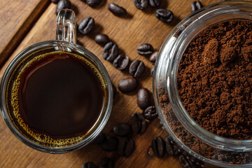 Xícara de café com vidro cheio de pó de café e grãos espalhados sobre a mesa de madeira.