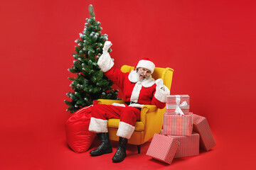 Joyful Santa Claus man in Christmas hat suit coat sit in armchair with fir tree presents gifts clenching fists like winner isolated on red background. Happy New Year celebration merry holiday concept.
