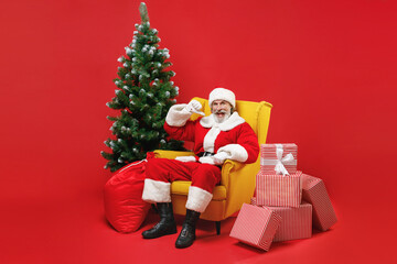 Excited Santa Claus man in Christmas hat suit sit in armchair with fir tree presents gifts point thumb on himself isolated on red background studio. Happy New Year celebration merry holiday concept.