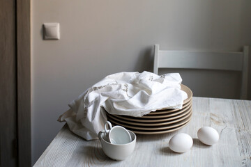 a stack of plates with an embroidered crumpled napkin, two eggs and two empty tea cups on a gray wooden kitchen table
