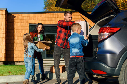 Joyful Family Helping Each Other To Unload Auto's Trunk From Carton Boxes And Suitcases During Moving To New Apartment