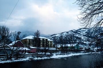 Riverside with houses, winter mountains, trees and a lot of snow. Winter background with blue sky