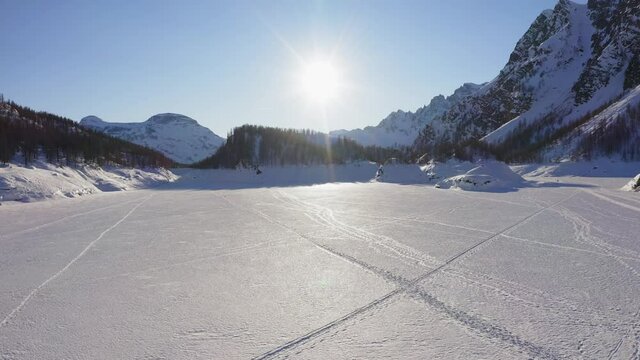 aerial over iced lake covered in snow on mountain valley in sunny winter day.Europe Alps mountains outdoor establisher.4k drone forward flight establishing shot. Sun flare