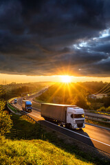 Trucks and delivery vans driving on the asphalt highway in forested landscape in the golden rays of the sunset with dark cloud - obrazy, fototapety, plakaty