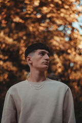 Hombre joven modelo en parque con arboles con hojas marrones. Ropa clara y marron 