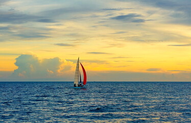 Yacht at sea at sunset.
Red sail. Orange sky.
Two people are driving the boat.