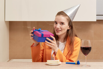 A happy young woman holds a gift in a bright box. On the head of a festive cap of silver color. A glass of red wine is on the table.