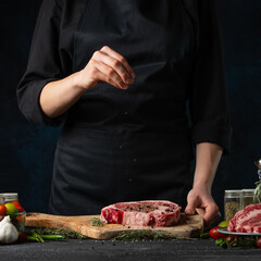 Professional chef in black uniform salts raw steak on wooden chopped board. Backstage of preparing grilled pork meat at restaurant kitchen on dark blue background. Food concept. Frozen motion.