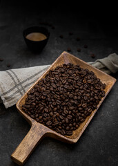 Inclined view of a wooden shovel with a great amount of roasted coffee beans on top of it. A stone background with some isolated coffee beans and a coffee cup are next to the shovel.