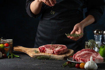 Close-up view of professional chef in black pours rosemary on raw steak on wooden chopped board. Backstage of preparing grilled pork meat at restaurant kitchen on dark blue background. Frozen motion.