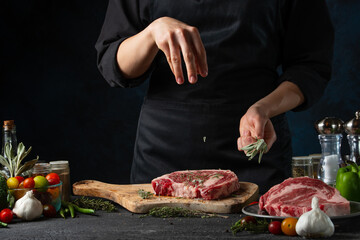 Close-up view of chef pours rosemary on raw steak on wooden chopped board. Backstage of preparing grilled pork meat at restaurant kitchen on dark blue background. Frozen motion. Close-up view.