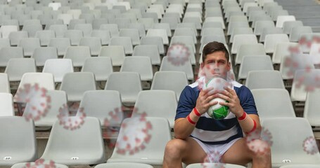 Covid-19 cells against male rugby player holding rugby ball and sitting in the stands of stadium - Powered by Adobe