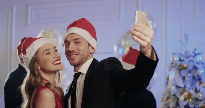 Young handsome man in suit with hat of Santa Claus takes selfie with beautiful woman i a red dress at corporate party.