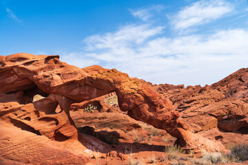 Fototapeta premium A natural sandstone arch in a red desert landscape under a clear blue sky with scattered clouds.