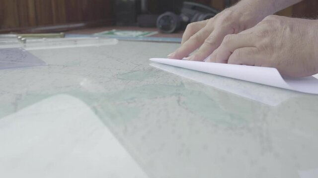 On a table with a map below the glass, a white paper is being folded into a boat like figure and shape.