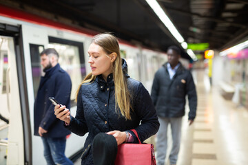 Portrait of female passenger waiting for train on subway platform and reading timetable in phone